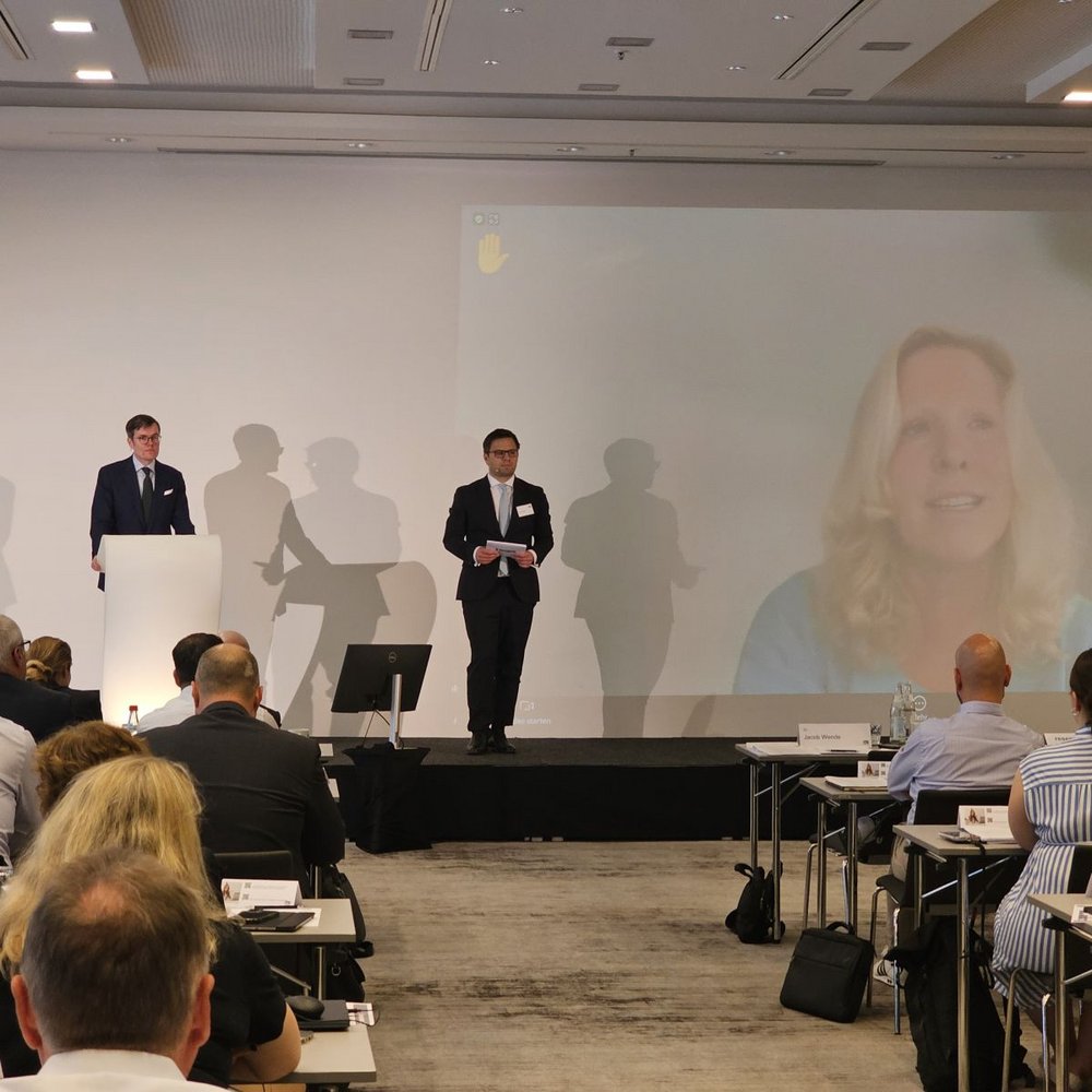 Zwei Referierende moderieren einen Programmpunkt auf der Bühne, während eine Referentin digital über eine große Videowand zugeschaltet ist.
