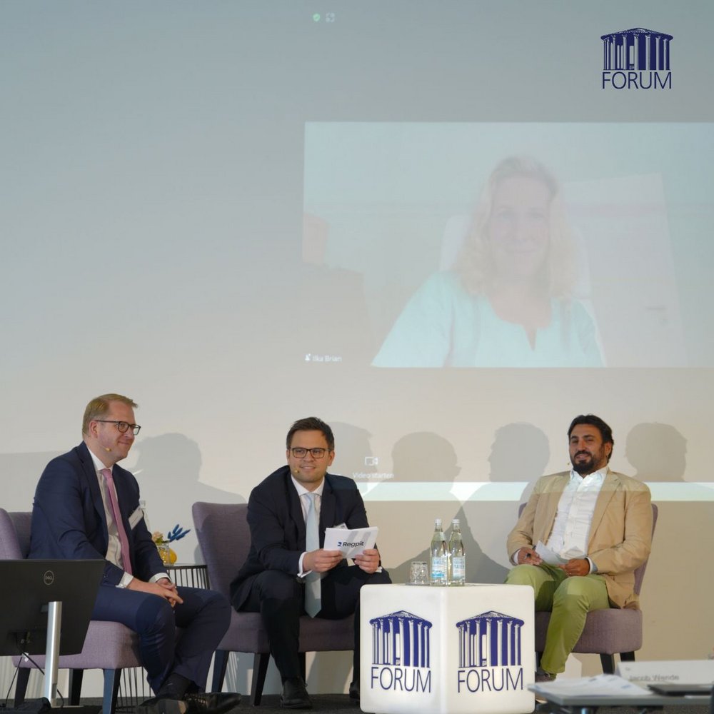 Drei Referierende sitzen auf der Bühne und diskutieren mit einer weiteren Referentin, die live per Videoübertragung zugeschaltet ist.