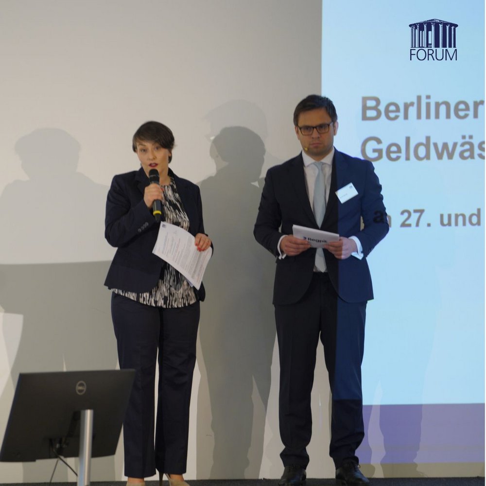 Die Konferenzmanagerin und der Tagungsleiter stehen gemeinsam auf der Bühne und eröffnen die Berliner Geldwäsche-Tagung vor dem Publikum im Saal.